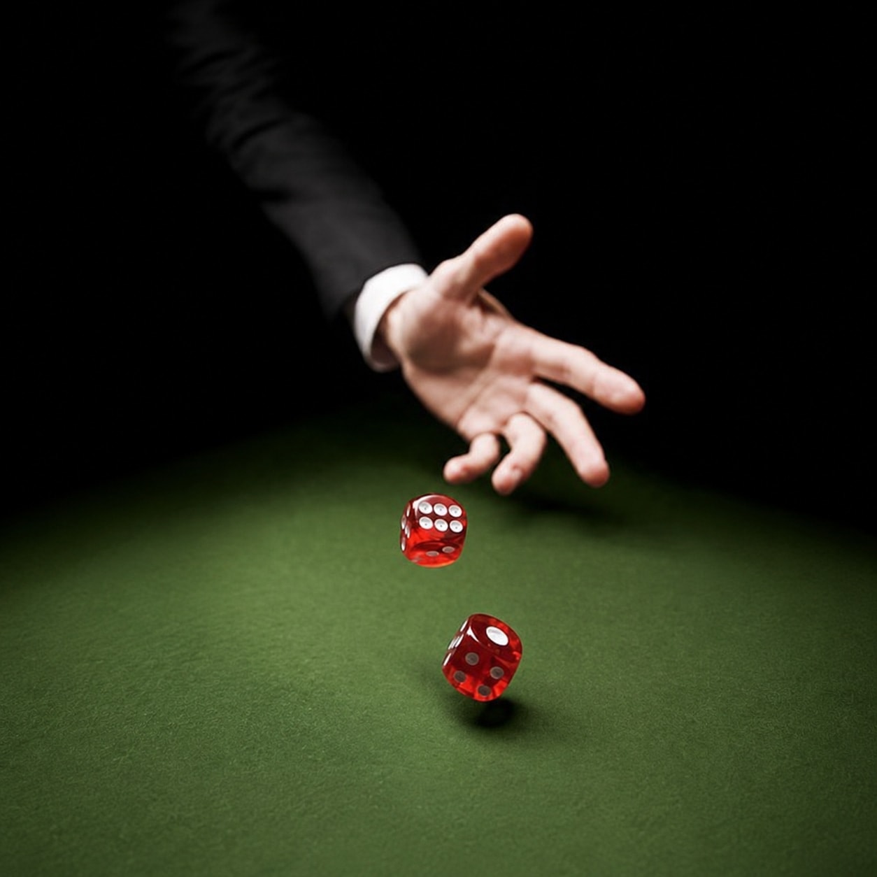 A hand in a dark suit throwing two red dice across a green casino felt table, lit from above against a black background