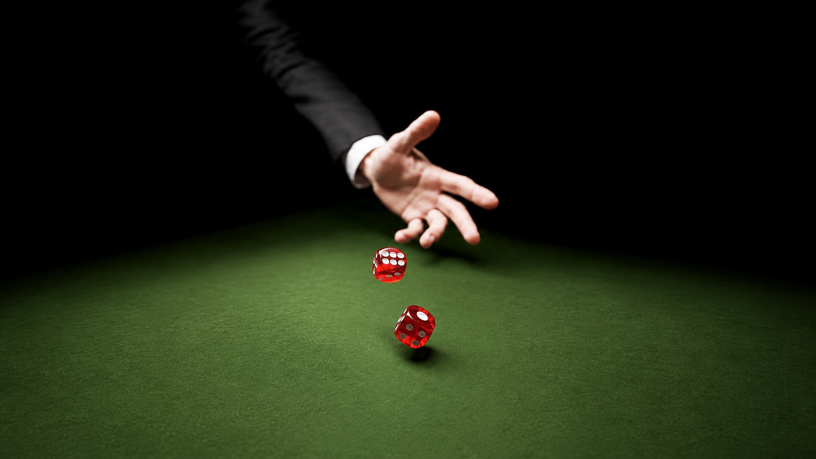 A hand in a dark suit throwing two red dice across a green casino felt table, lit from above against a black background