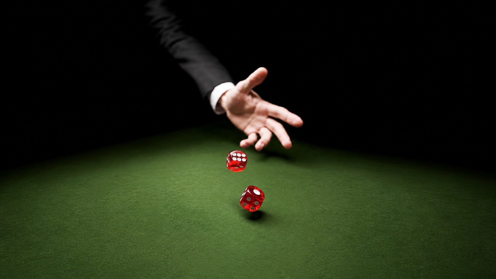 A hand in a dark suit throwing two red dice across a green casino felt table, lit from above against a black background